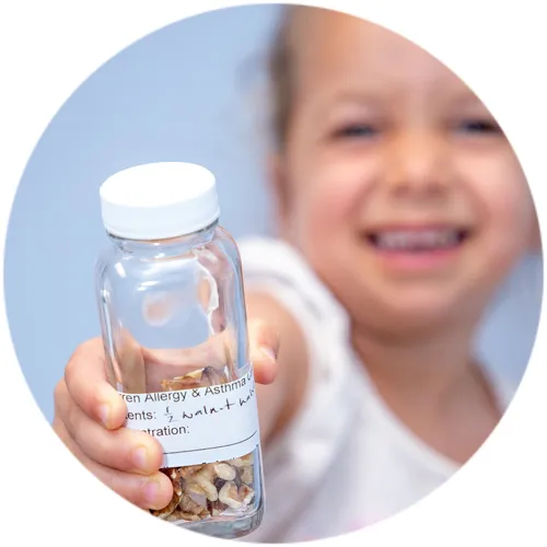 Smiling young girl holding up a labeled Warren Allergy & Asthma Care dosing bottle of walnut pieces